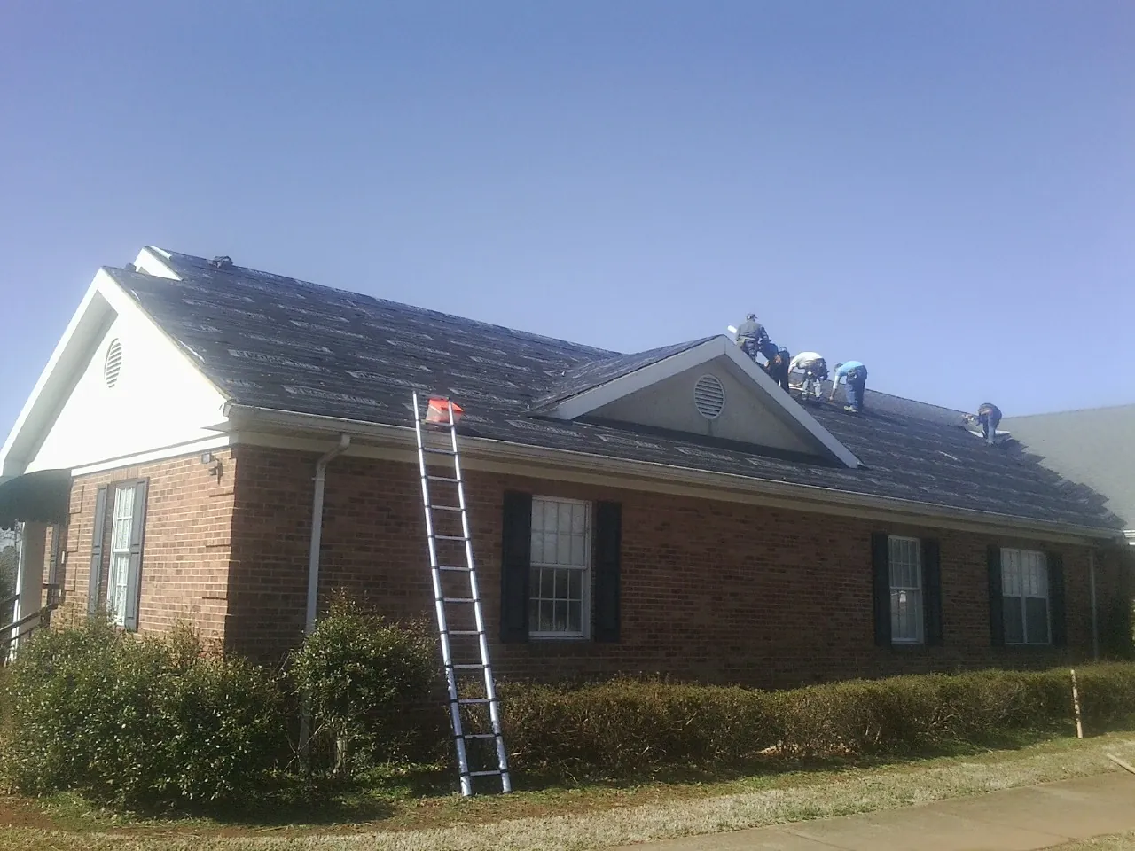 Crew tearing shingles off a brick ranch roof with a ladder set — Emergency Roof Repair work by our crew in Bayport